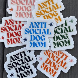 May include: A set of ten stickers with the text "Anti Social Dog Mom" in various colors and fonts. The stickers are on a wooden surface.