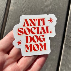 A different angle of a hand holding the red Anti Social Dog Mom vinyl waterproof sticker against a grey wood background, giving a better sense of scale. The sticker is perfect for the introverted dog mom in your life.