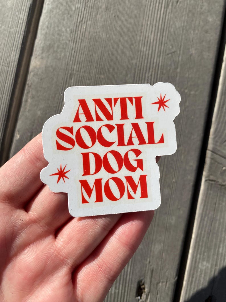 A different angle of a hand holding the red Anti Social Dog Mom vinyl waterproof sticker against a grey wood background, giving a better sense of scale. The sticker is perfect for the introverted dog mom in your life.