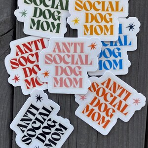 May include: A set of eight stickers with the text "Anti Social Dog Mom" in various colors and fonts. The stickers are on a wooden surface.