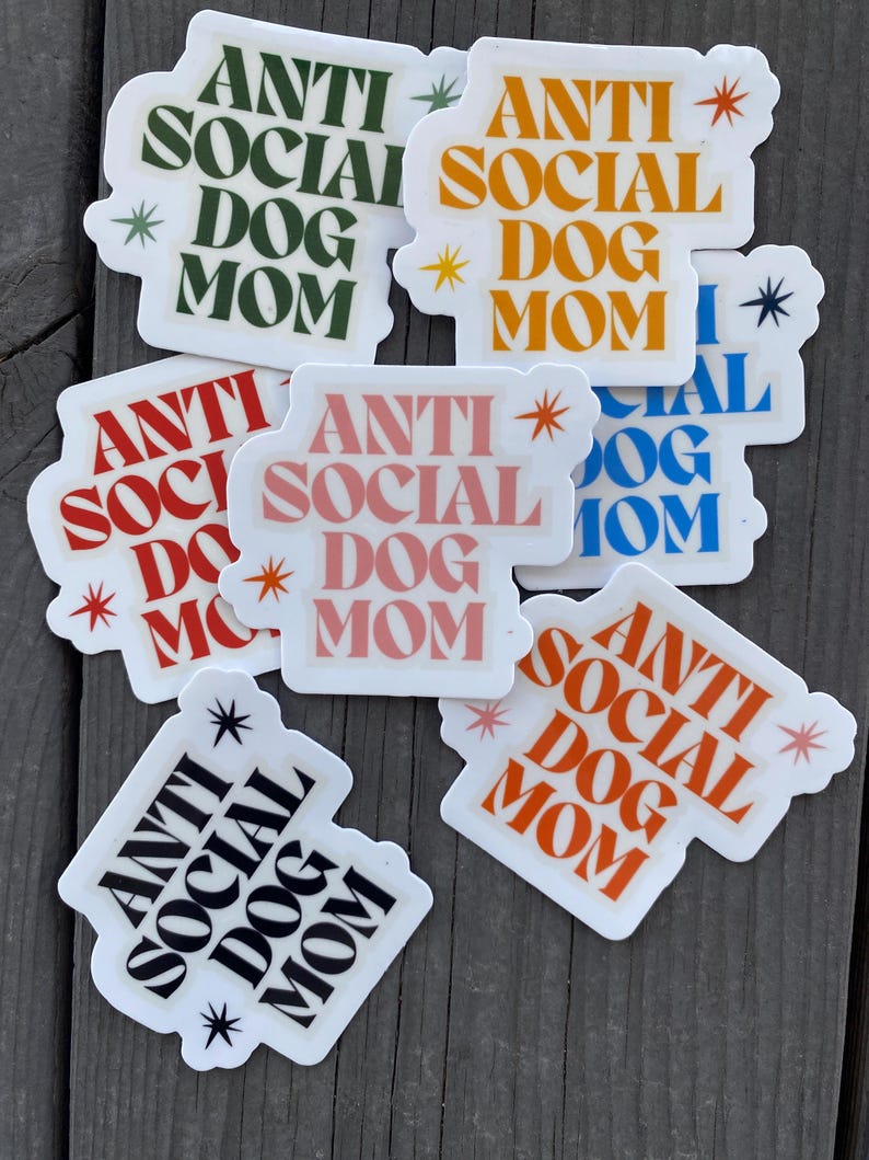 May include: A set of eight stickers with the text "Anti Social Dog Mom" in various colors and fonts. The stickers are on a wooden surface.