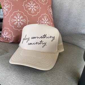 Tan “Play Something Country” Otto foam trucker hat with dark brown lettering sitting on porch rocking chair next to coral pillow, rustic country farmhouse style outdoor decor.