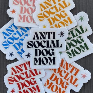 May include: A set of eight stickers with the text "Anti Social Dog Mom" in various colors and fonts. The stickers are on a wooden surface.
