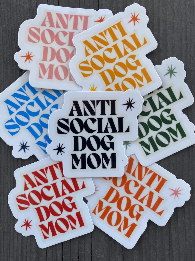 May include: A set of eight stickers with the text "Anti Social Dog Mom" in various colors and fonts. The stickers are on a wooden surface.