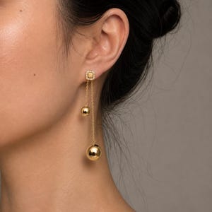 以下が含まれることがあります： 小さな白い石で縁取られた正方形のスタッドが付いたゴールドトーンのドロップイヤリング。スタッドから2本の繊細なゴールドチェーンが垂れ下がり、それぞれ異なるサイズの磨かれたゴールドの球体で終わる、レイヤード感のあるデザイン。