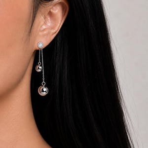 以下が含まれることがあります： 中央に白い石を配し、周囲を小さな透明なクリスタルで囲んだ小さな円形のスタッドが付いたシルバー調のドロップピアス。スタッドからは2本の繊細なシルバーチェーンが垂れ下がり、それぞれの先端に滑らかで光沢のあるシルバー調の球体ビーズが付いています。