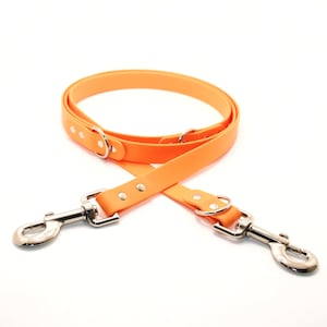 May include: Vibrant orange dog lead with two metal clasp attachments. The lead is made of a flexible material and features silver-coloured metal hardware, including rivets and D-rings. Designed for dual dog walking.