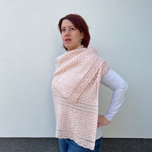 May include: A pink crocheted shawl with a delicate openwork pattern. The shawl is draped around a person's shoulders and is worn with a white long-sleeved shirt and blue jeans.