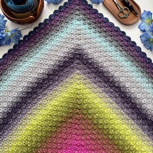 May include: A triangular crochet shawl featuring a gradient color pattern that transitions from vibrant pink and lime green at the point to deep purple, grey, and light blue at the wide edge. The shawl has a textured shell stitch and a scalloped border.