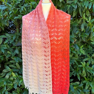 Puede incluir: Un chal de ganchillo con un degradado de melocotón, coral y blanco. El chal tiene un borde festoneado y está decorado con pequeñas borlas.