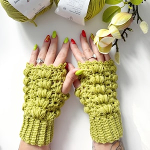 May include: A pair of lime green crochet fingerless gloves. The gloves feature a textured floral design and a ribbed cuff. The gloves are displayed on a white surface with yarn and flowers in the background. The gloves are handmade.