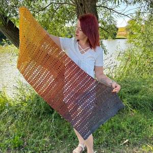 May include: A crocheted shawl with a gradient of colors from yellow to orange to red to blue. The shawl is held by a person standing in front of a lake.