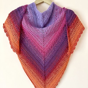 Puede incluir: Un chal de ganchillo en un degradado de naranja, rojo, rosa y morado. El chal está colgado en una percha de madera.