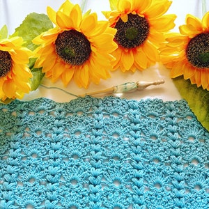 May include: A close-up of a crocheted blue blanket with four yellow sunflowers on top. A crochet hook is lying on the blanket.