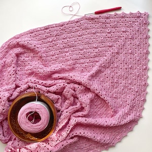 May include: A handmade pink crochet blanket with a textured pattern and scalloped edges. A wooden yarn bowl holds a ball of pink yarn. A red crochet hook and a heart-shaped yarn detail are also visible, suggesting a crafting project.
