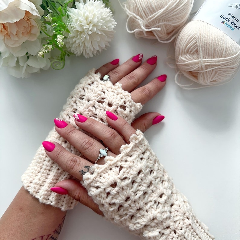 May include: A pair of crocheted fingerless gloves in a cream color. The gloves have a lacy pattern and are shown on a person's hands with pink nail polish.