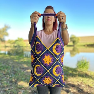 May include: A purple and yellow crocheted tote bag with a moon and star pattern. The bag is being held by a person.