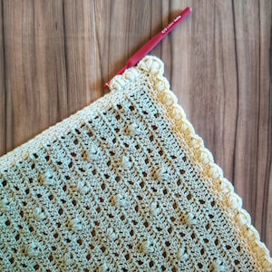 Könnte beinhalten: Nahaufnahme einer gehäkelten Decke mit einem gewellten Rand. Die Decke ist aus hellgrünem Garn gefertigt und hat ein strukturiertes Muster. Ein roter Häkelhaken ist in der oberen rechten Ecke des Bildes zu sehen.