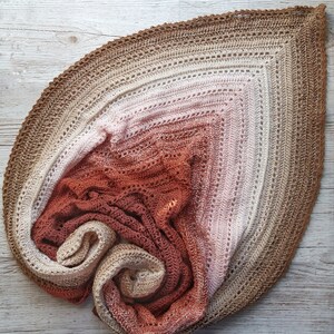 May include: A crocheted shawl in a gradient of brown, pink, and orange colors. The shawl is folded and arranged in a spiral shape.