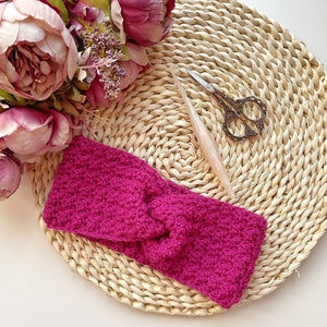 Puede incluir: Una diadema de crochet de color rosa brillante con un diseño anudado. La diadema está sobre una estera de paja tejida.