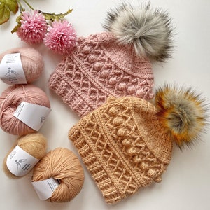 Peut inclure: Deux bonnets en crochet avec un motif de points texturés et des pompons. Un bonnet est rose et l'autre beige. Les bonnets sont entourés de pelotes de laine.