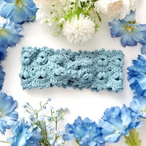 May include: A light blue crocheted headband with a floral pattern. The headband is made of soft yarn and features a textured design. The image is surrounded by blue and white flowers, creating a delicate and feminine aesthetic.