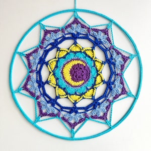 Op de afbeelding: Een gehaakte mandala wandhanger met een blauw, paars en geel kleurenschema. De mandala is rond en heeft een herhalend patroon van bloemen en bladeren.