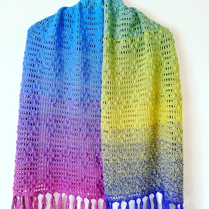 May include: A rainbow ombre crochet shawl with fringe on the bottom. The shawl is made with a light blue, green, yellow, and purple yarn.