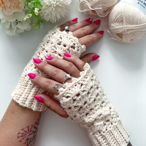 May include: A pair of crocheted fingerless gloves in a cream color. The gloves have a lacy pattern and are shown on a person's hands with pink nail polish.