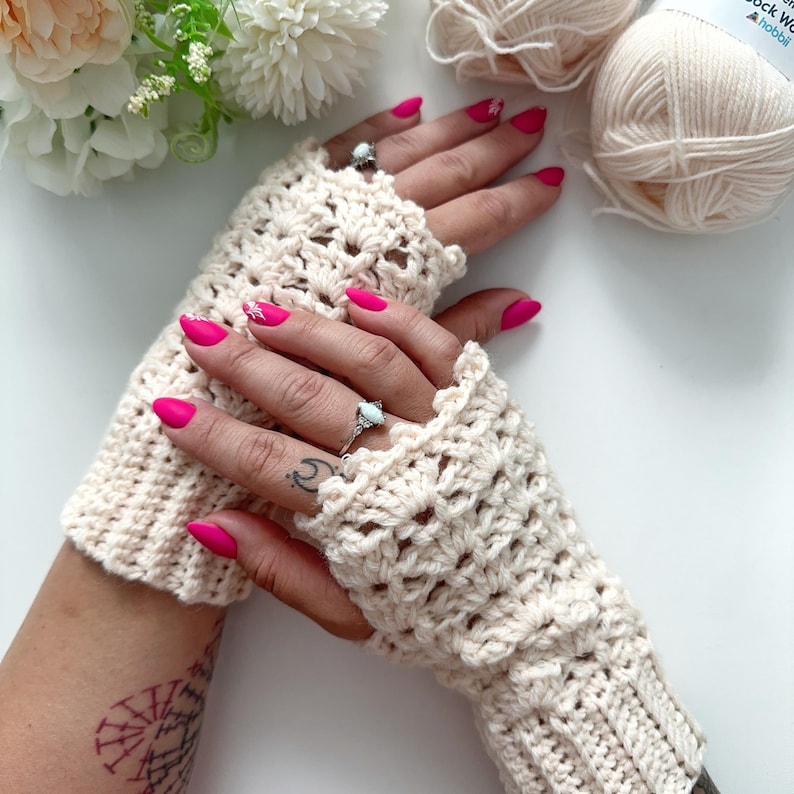 May include: A pair of crocheted fingerless gloves in a cream color. The gloves have a lacy pattern and are shown on a person's hands with pink nail polish.