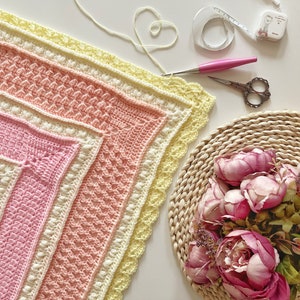 Può includere: Una copertina per neonati all'uncinetto in tonalità rosa, giallo e bianco con un bordo a festoni. La copertina è appoggiata su una superficie bianca con un paio di forbici, un metro a nastro e un gomitolo di lana.