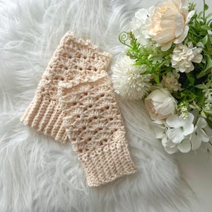 May include: A pair of crocheted fingerless gloves in a light cream color. The gloves have a delicate openwork pattern and are made with a soft yarn.