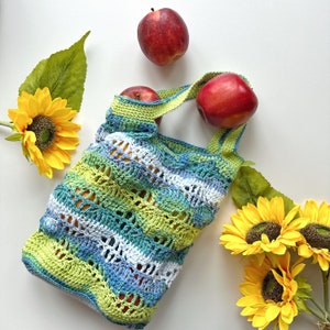 May include: A crocheted green, blue, and white tote bag with red apples inside. The bag is sitting on a white surface with yellow sunflowers.