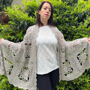 Może przedstawiać: Jasno szary szal wykonany na szydełku z ażurowym wzorem. Szal jest zarzucony na ramiona osoby ubranej w białą bluzkę i czarne spodnie.