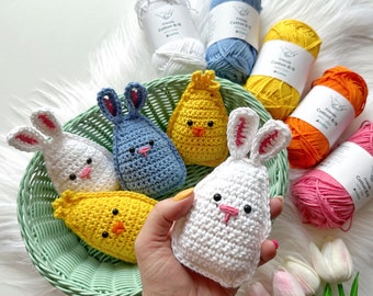 May include: A basket filled with crocheted Easter-themed animals, including a white bunny, a blue bunny, a yellow chick, and a yellow chick. The basket is surrounded by balls of yarn in various colors, including white, blue, yellow, orange, and pink.