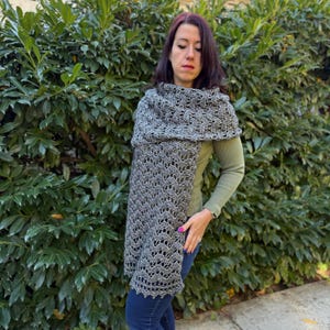 Peut inclure: Un châle en crochet gris avec un motif ajouré délicat. Le châle est drapé sur les épaules et descend sur le devant. La couleur est un mélange de gris et de noir. L'arrière-plan est un buisson vert.