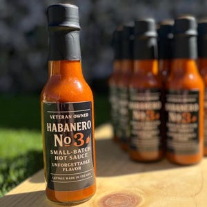 May include: A bottle of Habanero No. 3 small-batch hot sauce, with a black label and a black cap. The label reads "Veteran Owned" and "Unforgettable Flavor." Several other bottles are visible in the background, all on a wooden surface.