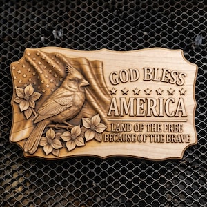 May include: Wooden plaque with a carved design featuring an American flag, a cardinal bird, and floral accents. The text "GOD BLESS AMERICA" and "LAND OF THE FREE BECAUSE OF THE BRAVE" are also carved into the wood.