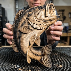 May include: A detailed wooden carving of a fish, featuring intricate scales and fins. The fish is a light brown color with darker brown accents, and is held up by a person. The carving is displayed against a blurred background.