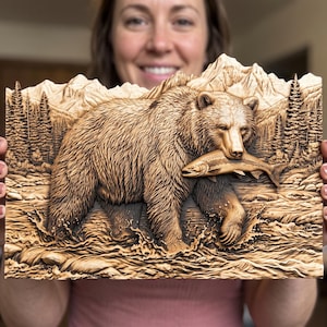 May include: A rectangular wooden wall plaque featuring a detailed relief carving of a grizzly bear walking through a river while holding a fish in its mouth. The background depicts a forest of pine trees and jagged mountain peaks in natural wood tones.