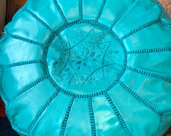 May include: A turquoise leather pouf with a star-shaped design in the center. The pouf has a stitched border and is made of a soft, supple leather.