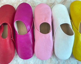 May include: Five slip-on slippers arranged in a row, featuring smooth leather construction with rounded toes and open heel designs. The colors from left to right are deep red, vibrant magenta, soft pastel pink, crisp white, and bright yellow.