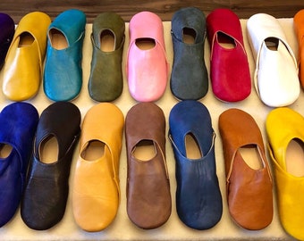 May include: A collection of sixteen slip-on slippers arranged in two rows. The slippers are made of solid-colored leather in various shades including purple, yellow, turquoise, olive green, pink, dark gray, red, white, orange, blue, brown, and fuchsia.