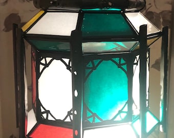 May include: A black metal lantern with stained glass panels in red, white, green, and yellow. The lantern has a decorative, cut-out design on the top and bottom.