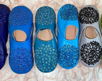 Puede incluir: Seis pares de zapatillas de cuero con intrincados diseños bordados. Las zapatillas son de varios tonos de azul, gris y verde. Cada zapatilla presenta un diseño circular con lentejuelas y cuentas.