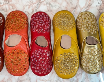 Puede incluir: Seis pares de zapatillas de cuero con intrincados diseños bordados. Las zapatillas son de varios colores, incluyendo marrón, naranja, rojo, amarillo y dorado. Cada zapatilla presenta un patrón único de lentejuelas y cuentas.