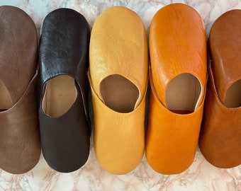 May include: Five leather slippers arranged in a row, showcasing a gradient of colours from left to right: light brown, dark brown, pale yellow, golden orange, and medium tan. Each slipper features a rounded toe and a simple slip-on design.