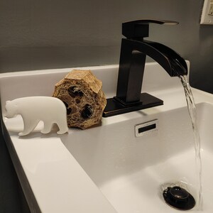 May include: A white ceramic bear figurine sits on the edge of a white porcelain sink. A brown loofah sponge sits behind the bear. A black faucet with a curved spout is turned on, and water is flowing into the sink.