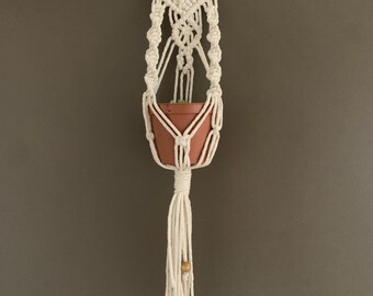 May include: White macrame plant hanger with a brown terracotta pot and wooden beads hanging from the bottom.