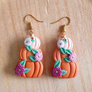 May include: A pair of gold dangle earrings featuring three stacked orange, peach, and pink pumpkins with green leaves and pink and white flowers.